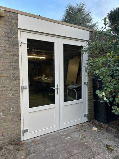 Kozijnplaatsing in Vlijmen Witte dubbele deuren met glazen panelen, gelegen in een stenen muur met groenblijvende planten.