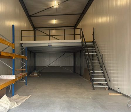 Lege opslagruimte met mezzanine en metalen trap, houten planken en betonnen vloer.