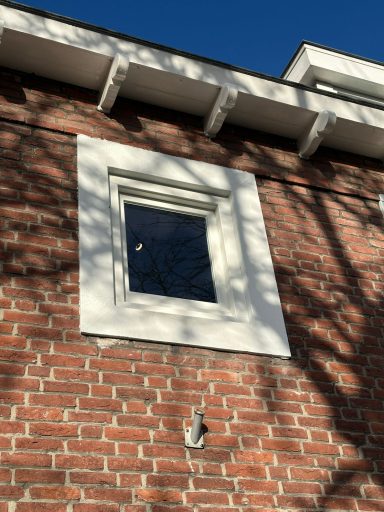 Kozijnplaatsing in 's-Hertogenbosch Raam met wit kozijn in een bakstenen muur, schaduw van bomen op de gevel.
