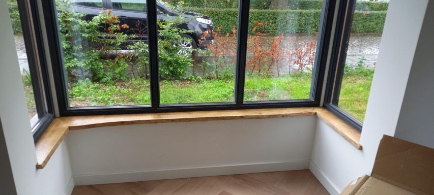 Vensterbanken in Maliskamp Hoekbank met lichtkleurige afwerking bij een raam, uitzicht op een groene tuin.